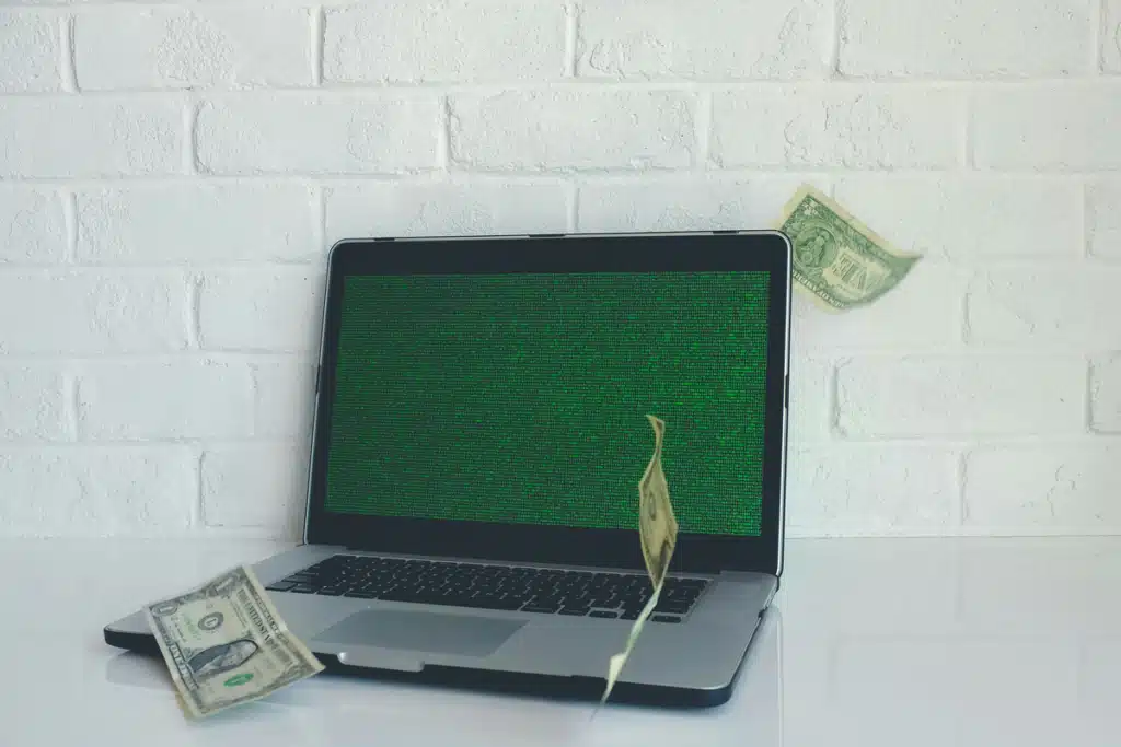 Cash money flying around a laptop on a white desk in front of a white brick wall.