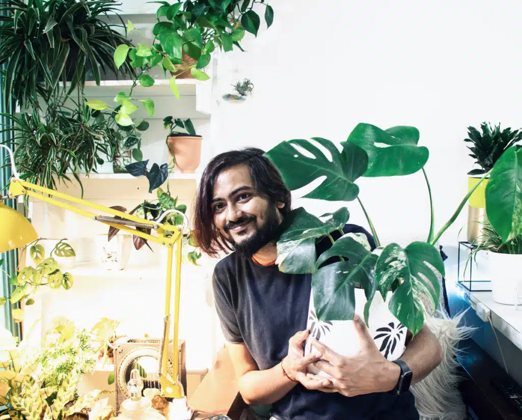 A guy is happily holding a potted plant. There is a cool yellow desk lamp behind him, and many other plants.