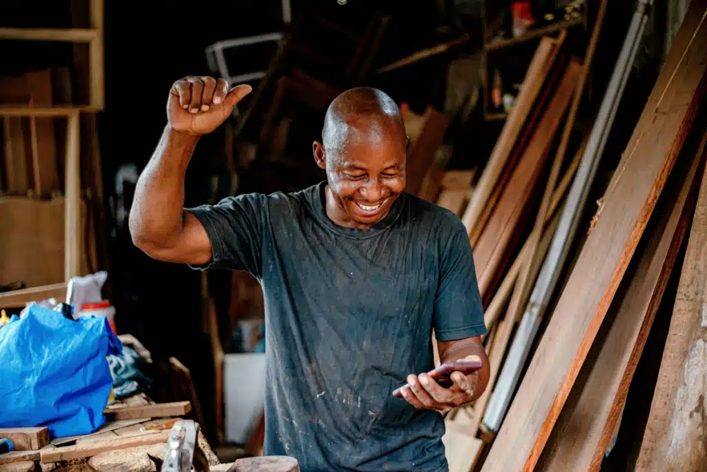 A small business owner is standing in his workshop. He looks at the mobile phone in his hand and smiles, throwing a hand in the air in celebration. He has found international shipping for small business on PostFromUS.