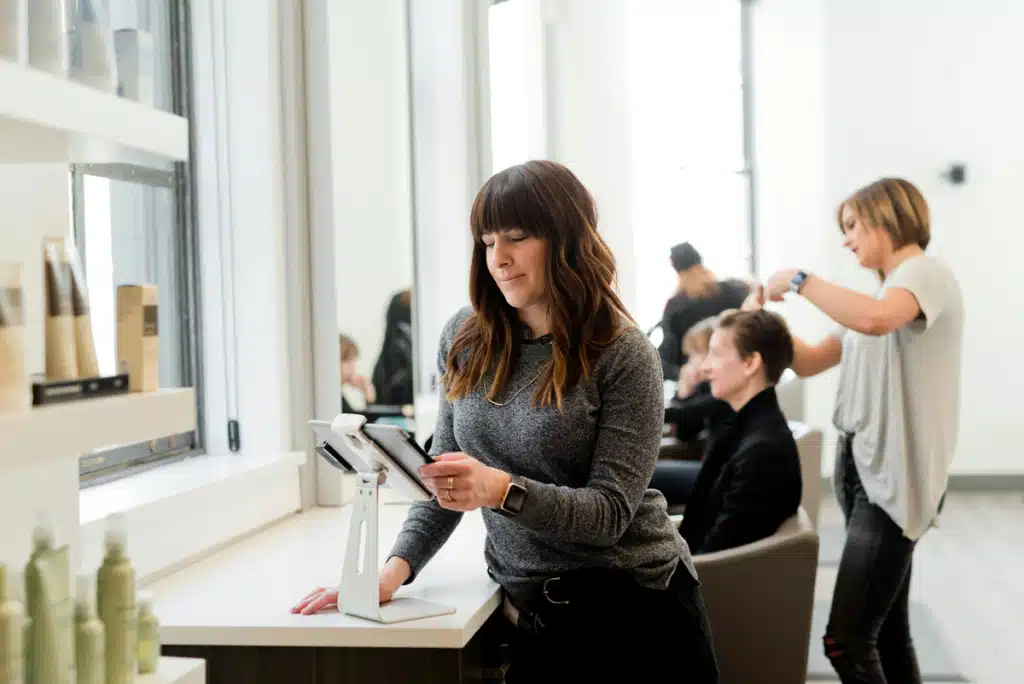 A woman is inside her hair salon, searching on her phone for how to get the best shipping for small business. This can help her sell products to more customers!