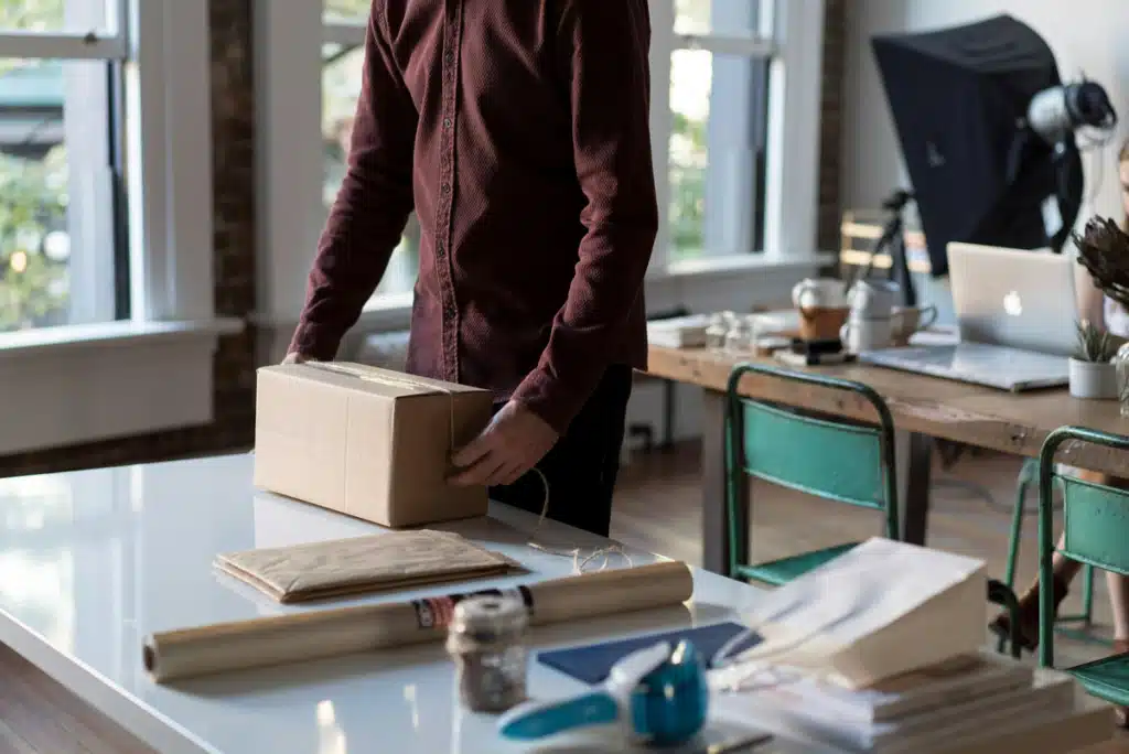 Someone is preparing their postage to Hawaii on a desk with various packaging materials around them like wrapping paper. There is a box in their hands. 