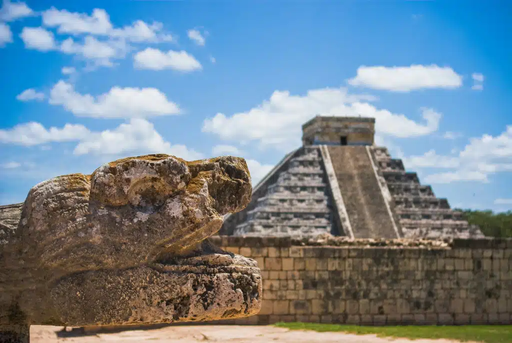 A Mexican ruin that tourists flock to, resembling a pyramid made of stone. A rock formation is in the foreground. The sky is blue and partially cloudy.