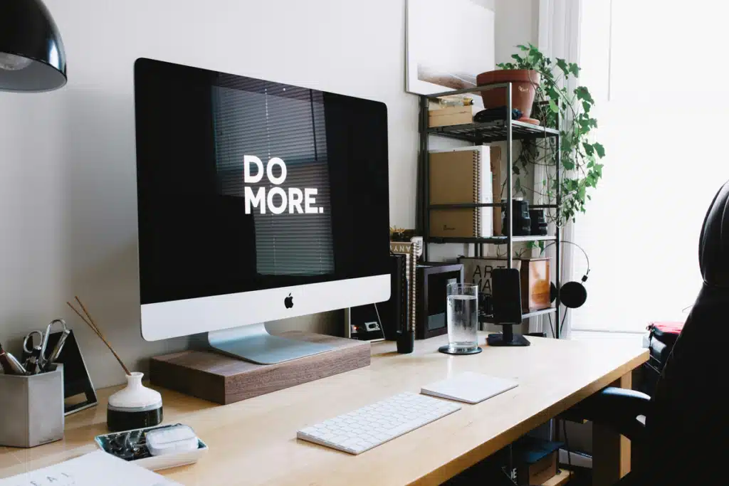 An iMac is set up on a desk with matching accessories. The screen says "DO MORE." in white text on a black background. Do more by signing up with PostFromUS to save when Shipping to New Zealand from USA.