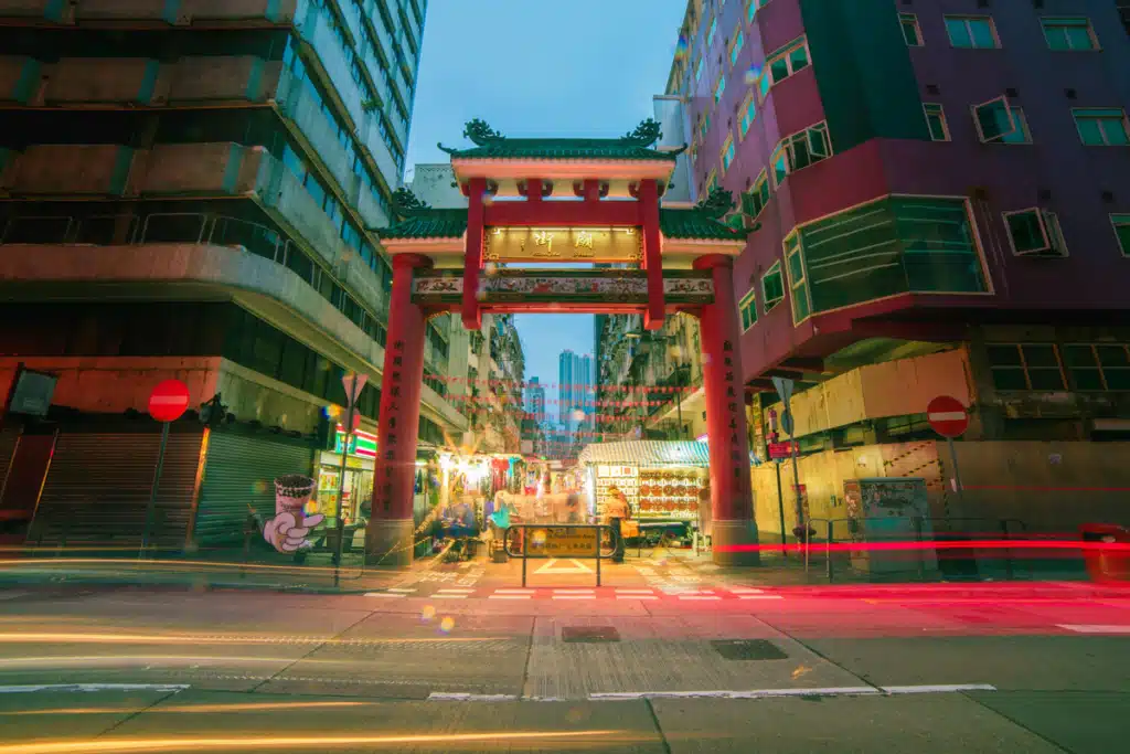 A red Chinese style city gate is over a road in between two skyscrapers. The sun is going down and the city lights are glowing.