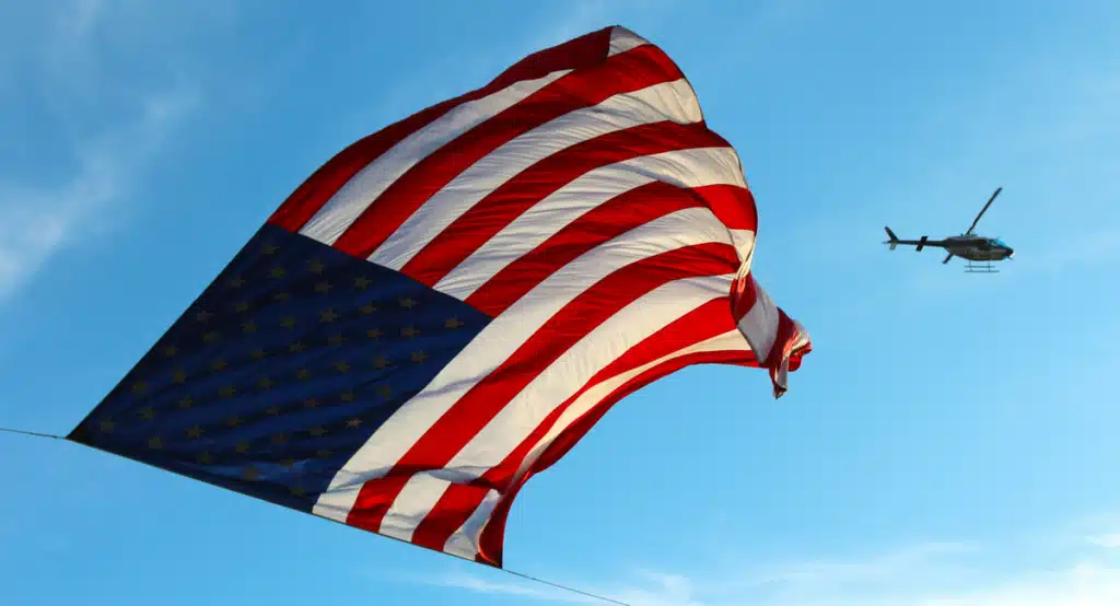 US flag flying in the air, a helicopter from the US military is flying high above it.