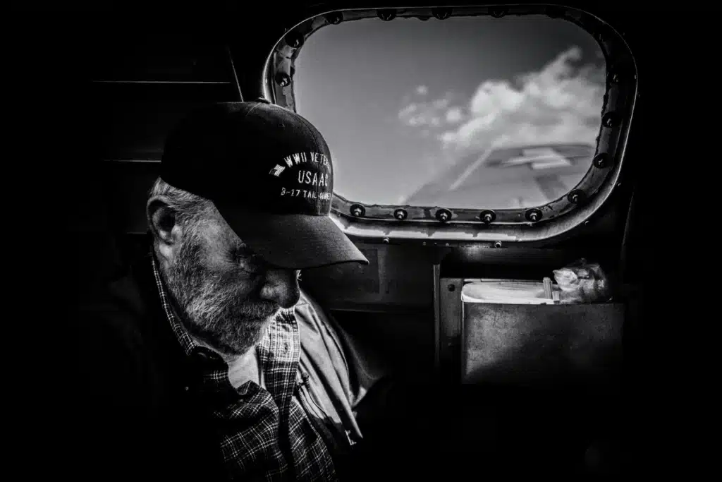 In greyscale, a man in the US military is inside a plane, waiting for his package to be delivered. He is wearing a veteran's hat, likely stationed in a reserve force.