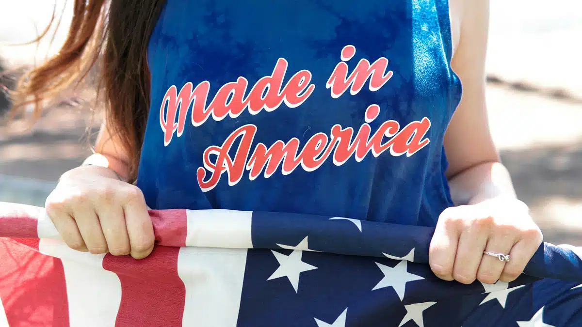 A woman is wearing a tank top that says 'Made in America' in red text over blue fabric. She's outside, holding a US flag. She's rolling it up because she's learning how to send mail to a US military base.