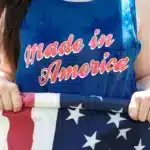 A woman is wearing a tank top that says 'Made in America' in red text over blue fabric. She's outside, holding a US flag. She's rolling it up because she's learning how to send mail to a US military base.