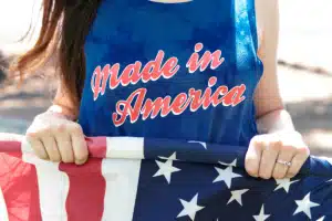 A woman is wearing a tank top that says 'Made in America' in red text over blue fabric. She's outside, holding a US flag. She's rolling it up because she's learning how to send mail to a US military base.