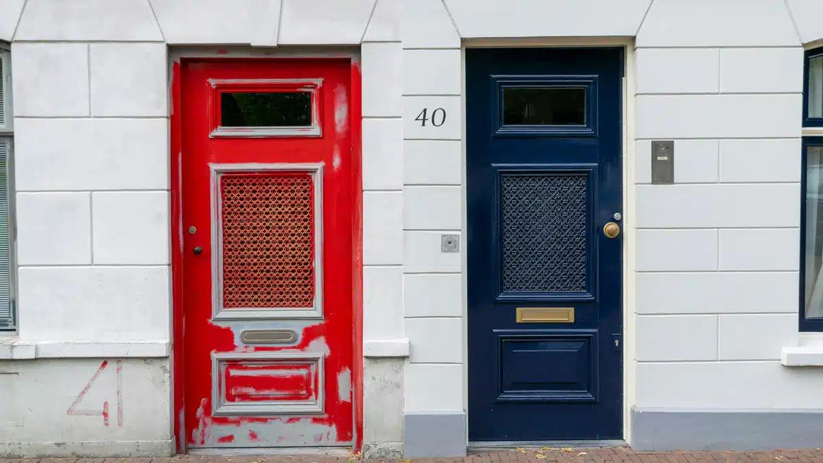 A white building in the Netherlands with a red door on the left, blue door on the right. Ship here from the US with PostFromUS.