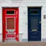 A white building in the Netherlands with a red door on the left, blue door on the right. Ship here from the US with PostFromUS.