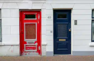 A white building in the Netherlands with a red door on the left, blue door on the right. Ship here from the US with PostFromUS.