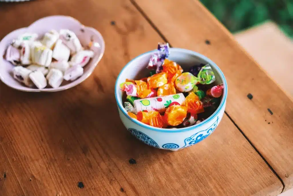 A pink scalloped edge candy dish, and a blue round candy dish with a darker blue pattern painted on it, are full of candy. They're on a wooden plank table top.