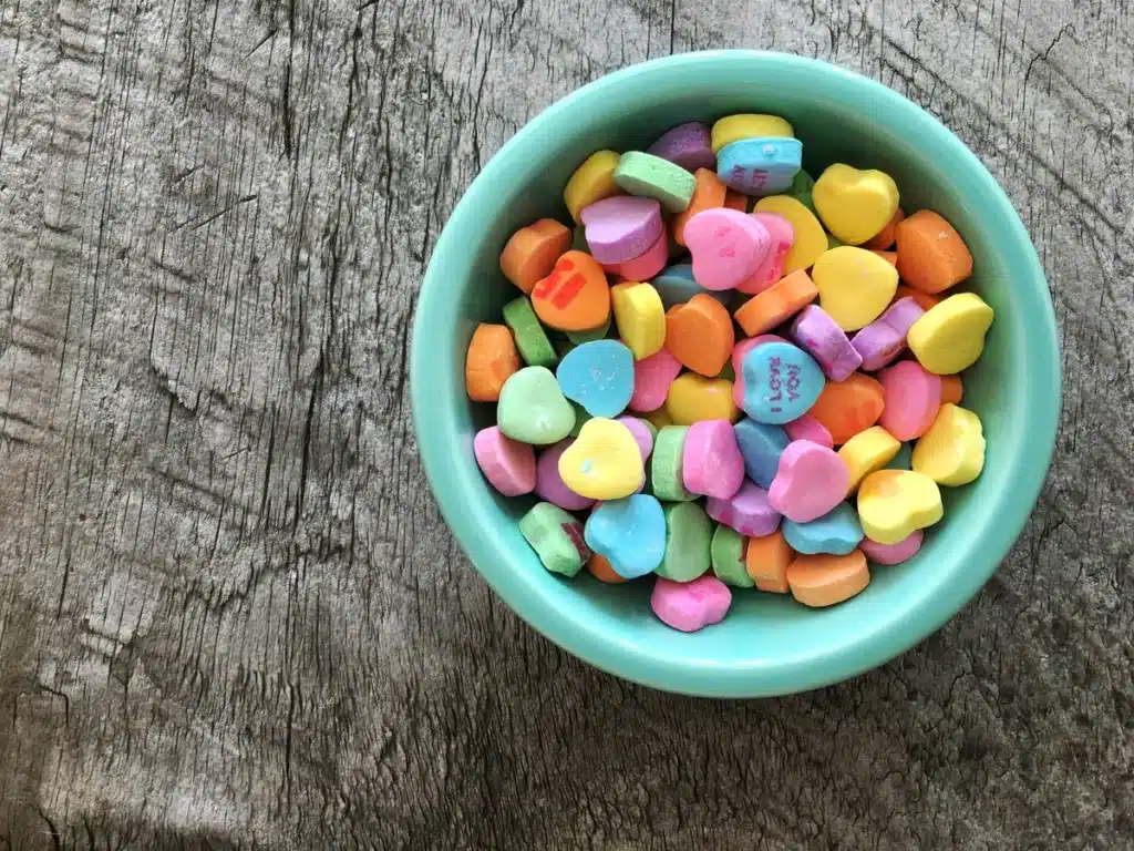 Valentine's Day love heart candies are in a unique teal-colored candy dish on top of a grey wooden background.