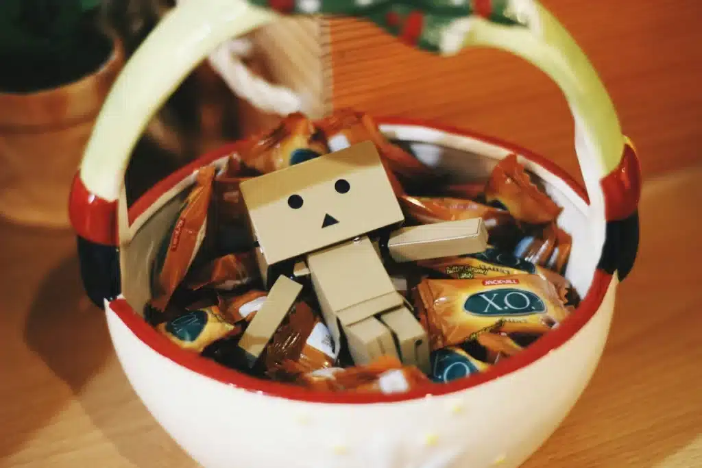 A round white unique Christmas candy dish with a handle, and some red features painted on it. Inside the bowl, there is candy and a toy robot.