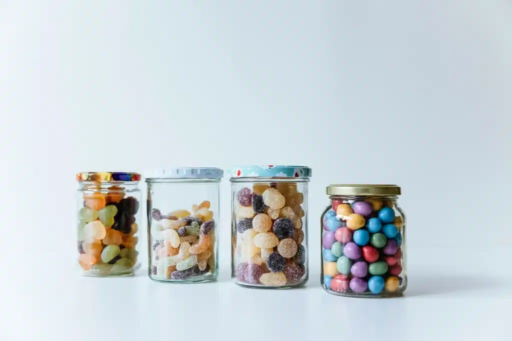 Four unique glass candy dishes with lids are lined up on a white surface in front of a white background, showing how full they are of different types of candies.