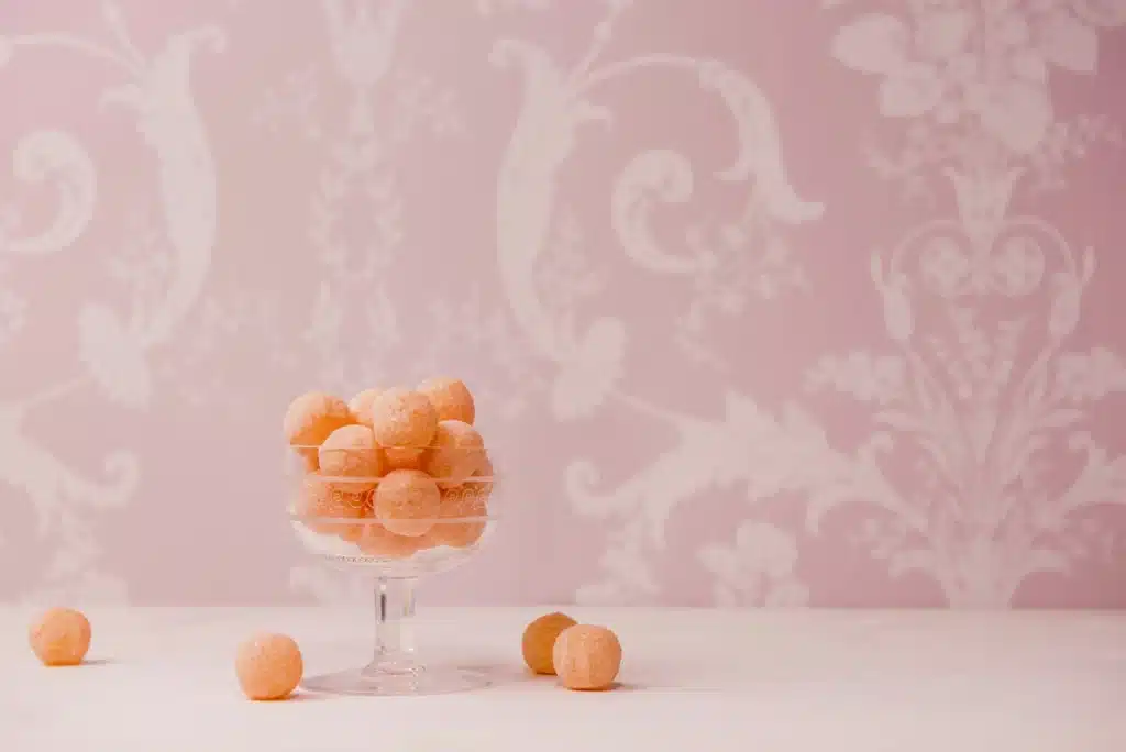 A unique glass candy dish is holding round orange candies in front of a pink antique wallpaper.